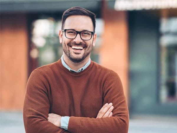 Professional man with glasses and crossed arms smiling outdoors in front of brick building.
