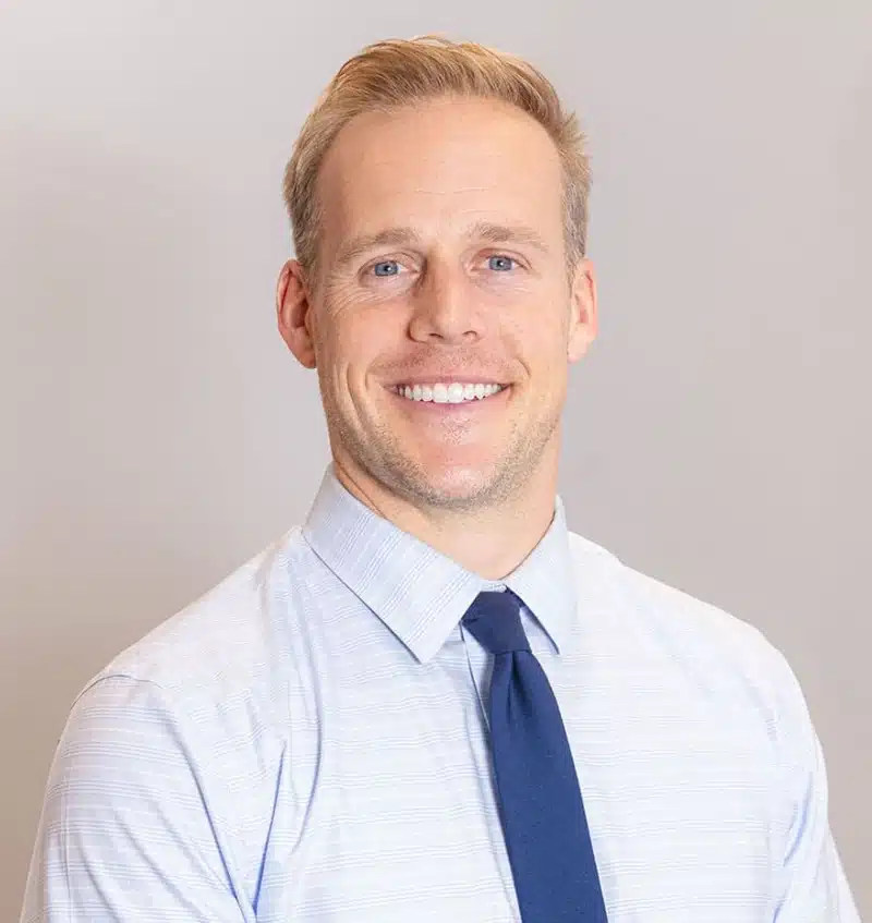 Professional headshot of smiling male dental professional in light blue shirt and navy tie